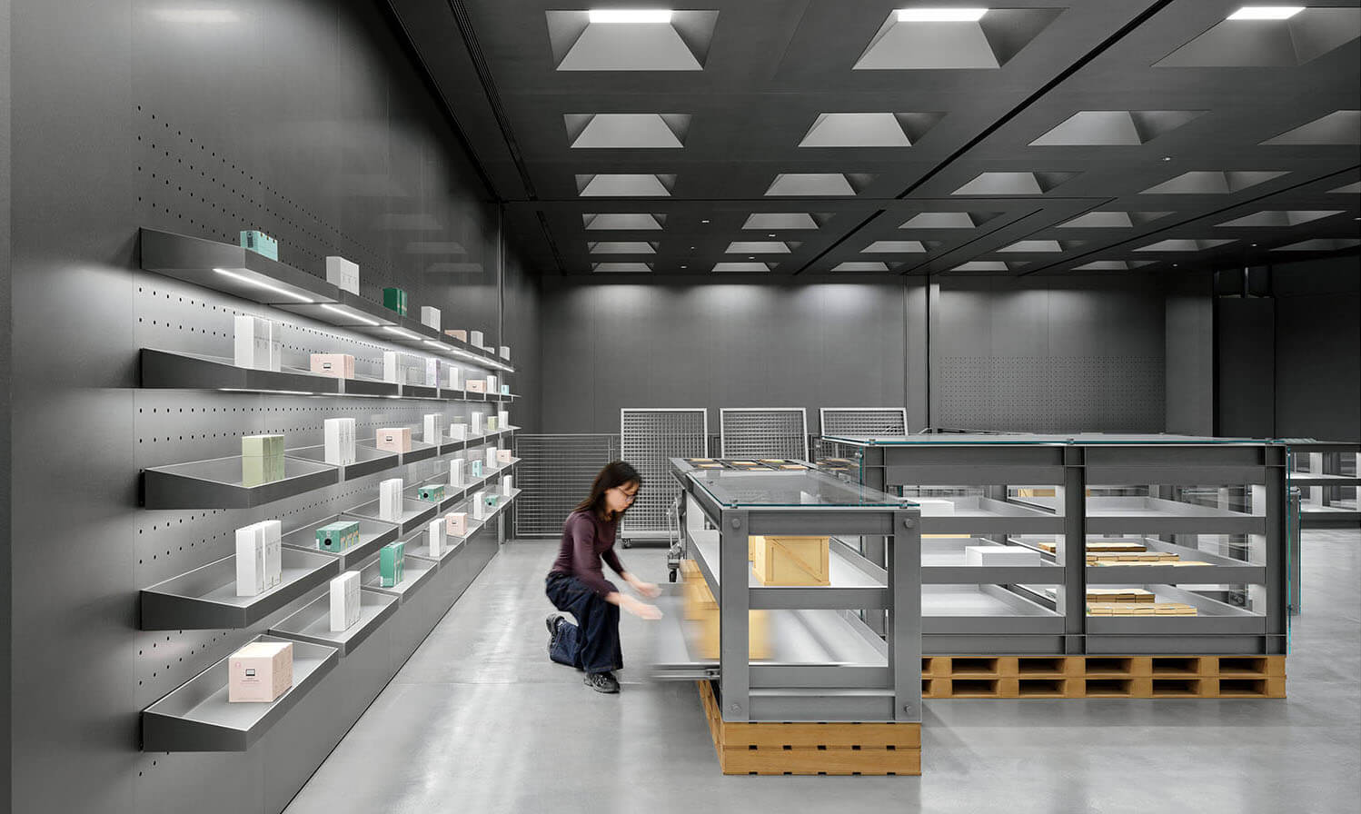 A person arranges items in a modern, minimalist store with metal shelves displaying small boxes and a gray color scheme.