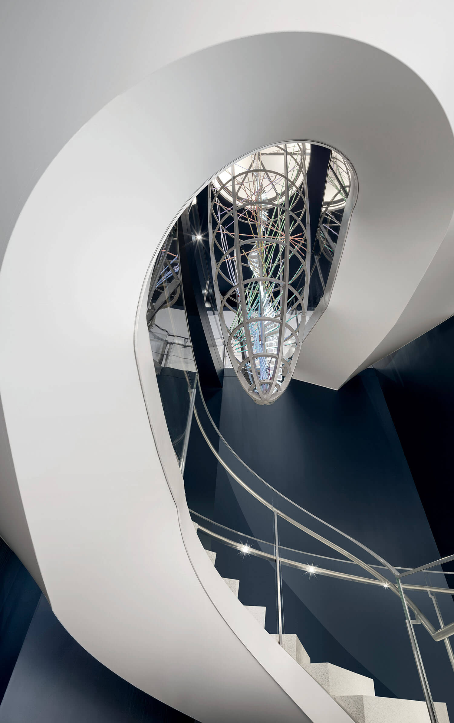 Curved white staircase with glass railing spirals upward beneath a geometric metal and glass light fixture, viewed from below.
