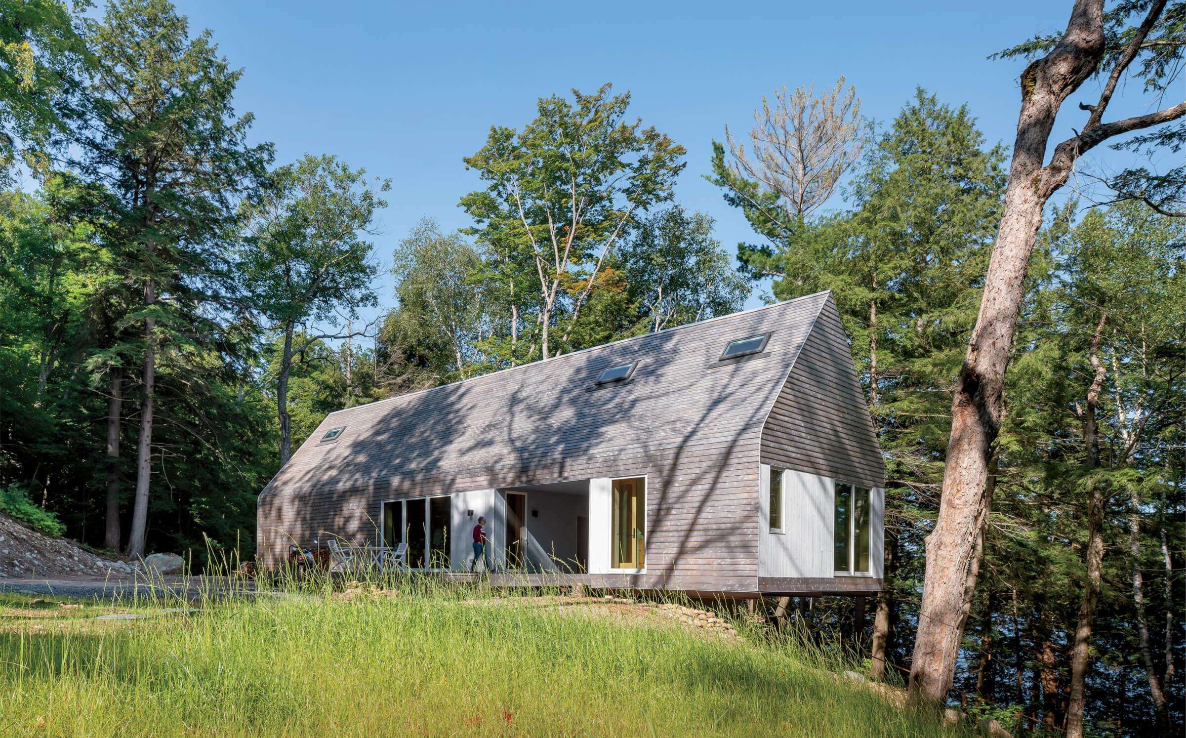 A modern, rectangular wooden house with large windows sits elevated among trees, surrounded by grass and overlooking a body of water.