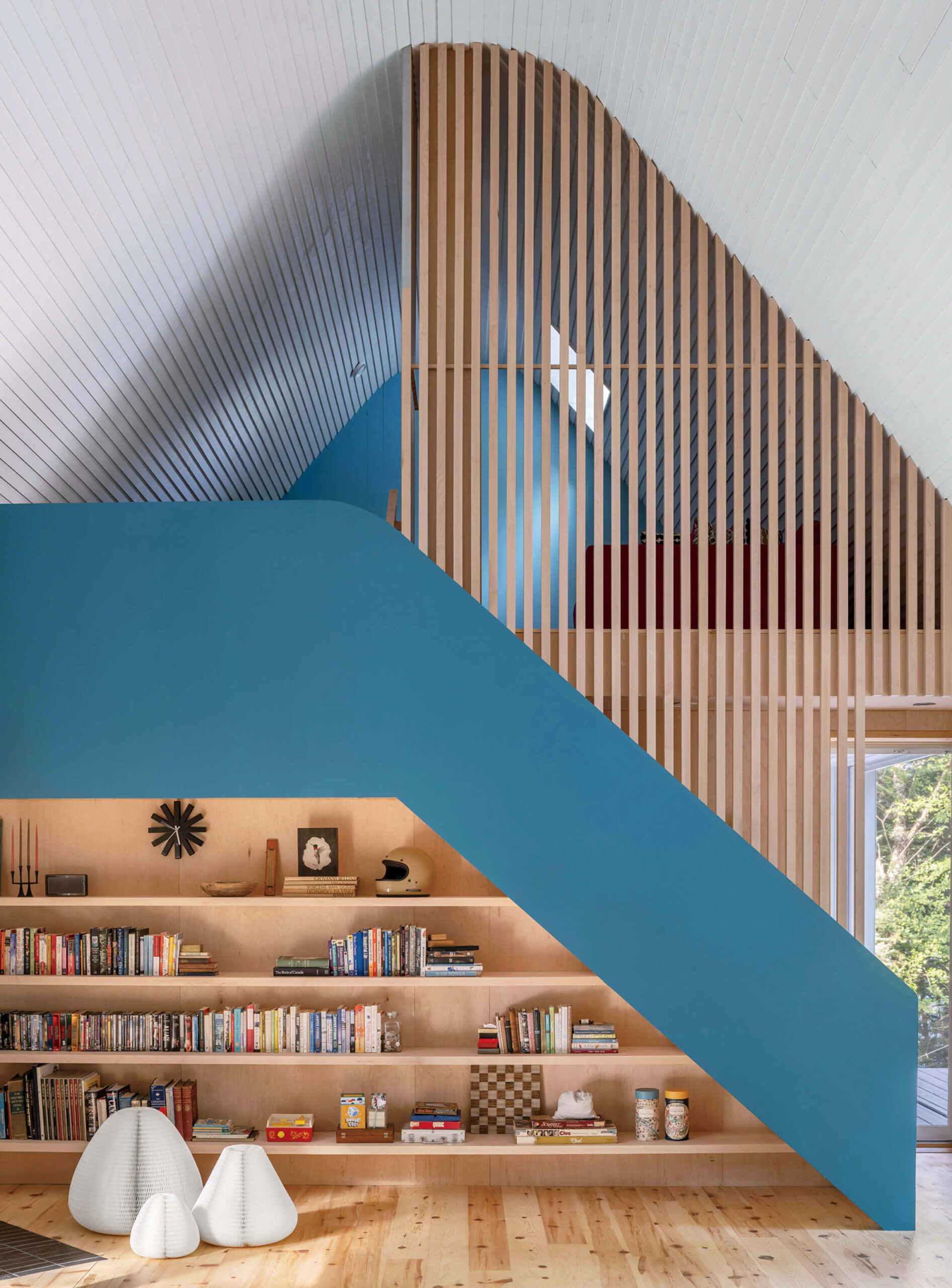 Modern interior with blue staircase, wooden slat wall, built-in bookshelf, and minimalist decor; natural light enters from side window.