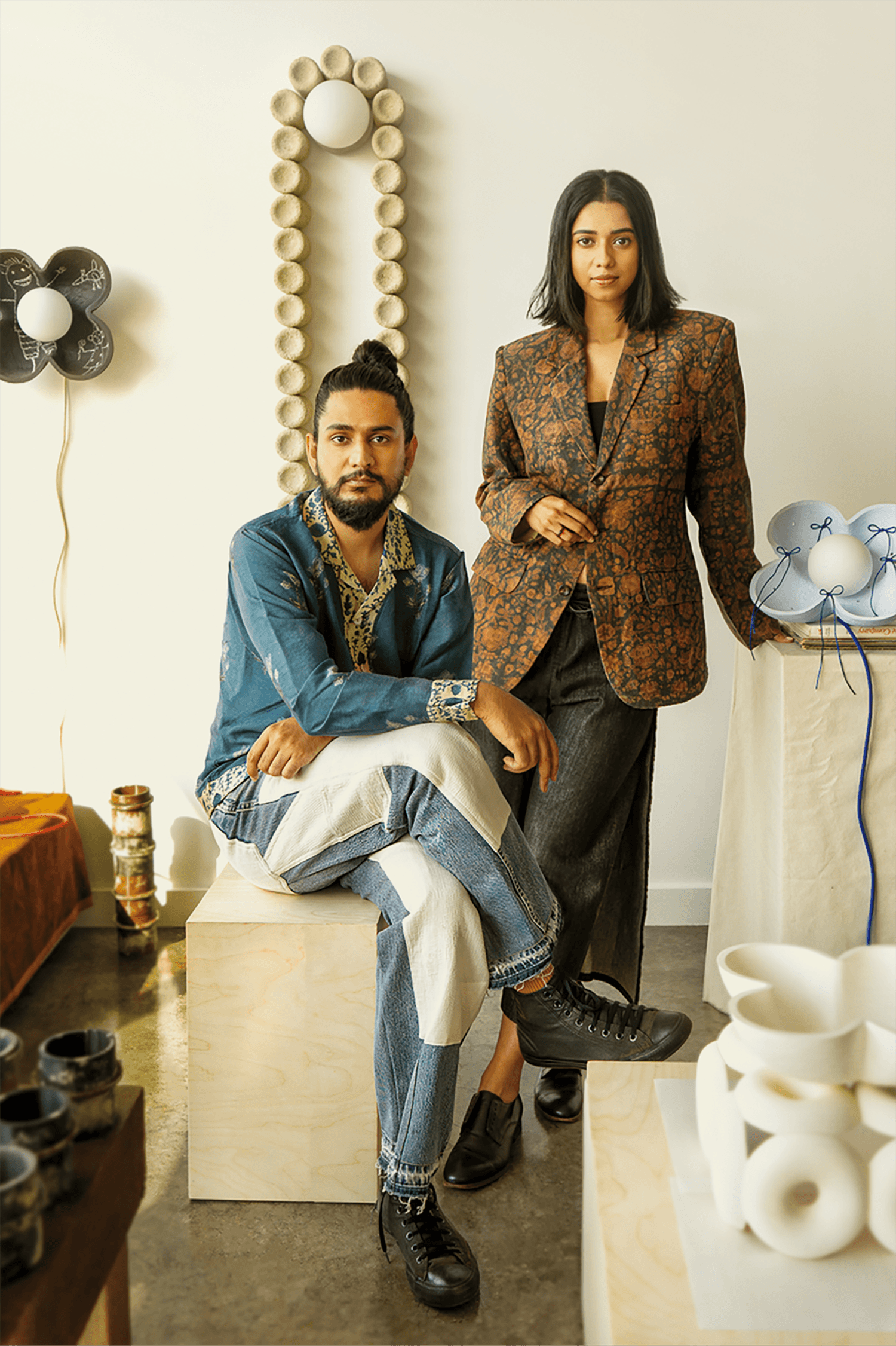 A man in a blue jacket and jeans sits on a wooden block next to a woman in a patterned blazer, standing in a room decorated with ceramics and modern lighting fixtures.