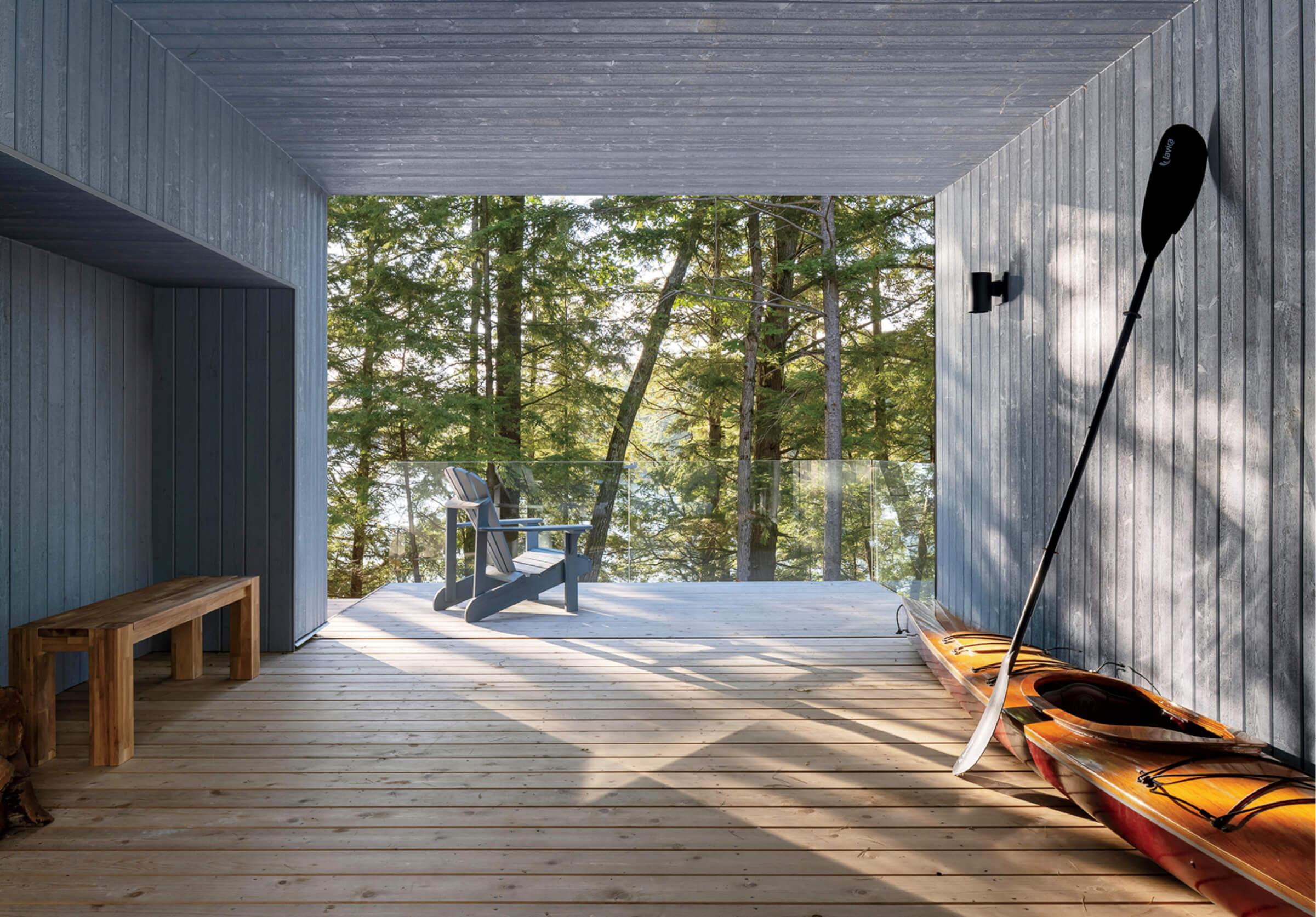 A wooden porch with a bench, a kayak, and a paddle faces a deck with a chair. The area overlooks trees and a distant body of water.
