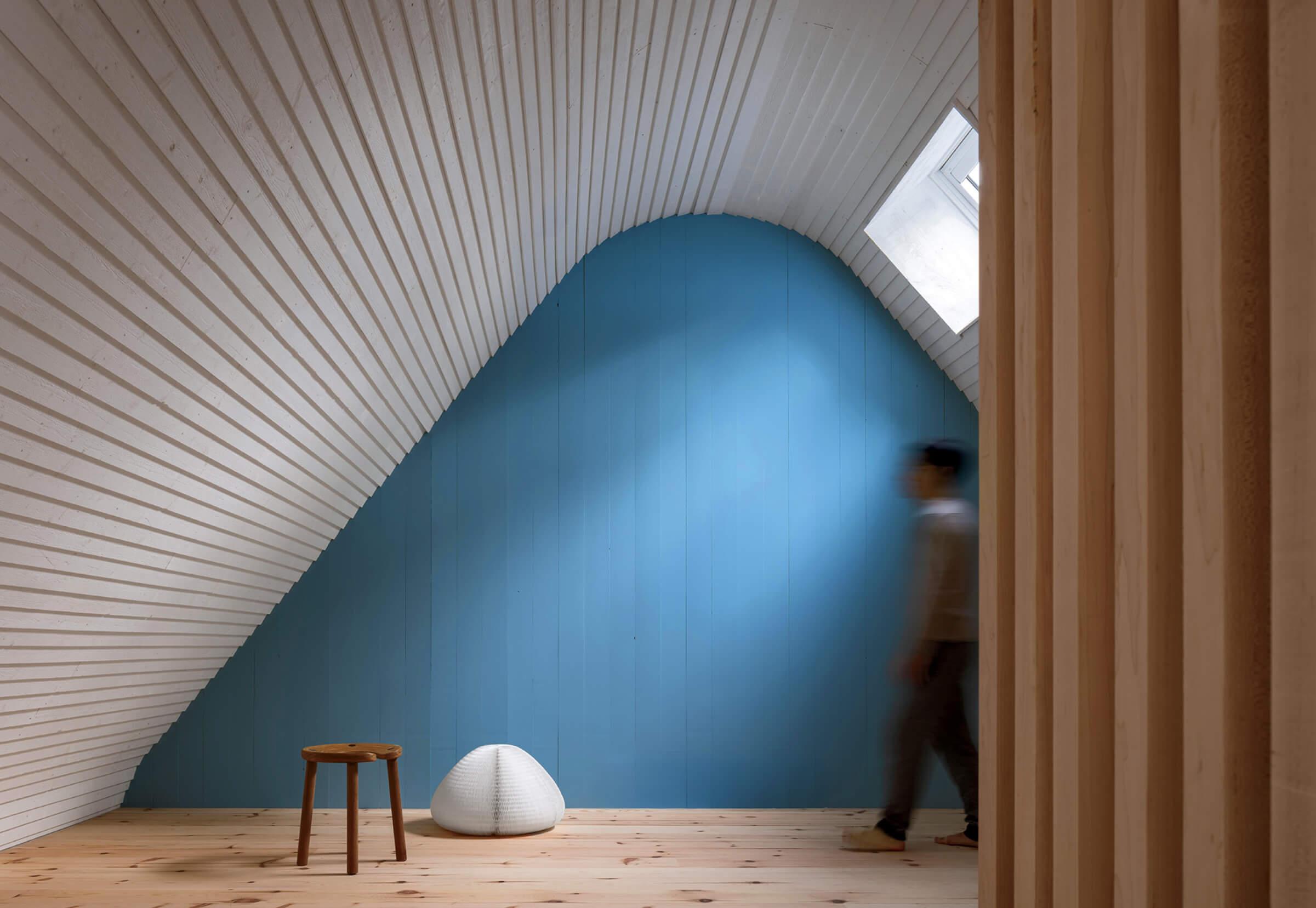 A person walks in a minimalist room with a curved white ceiling, blue accent wall, wooden floor, stool, a white object, and natural light from a skylight.