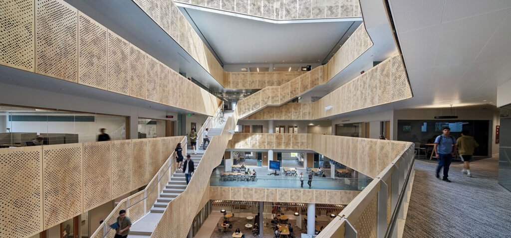 Open, modern building interior with multiple floors, angular staircases, perforated metal railings, and people walking or sitting in common areas.