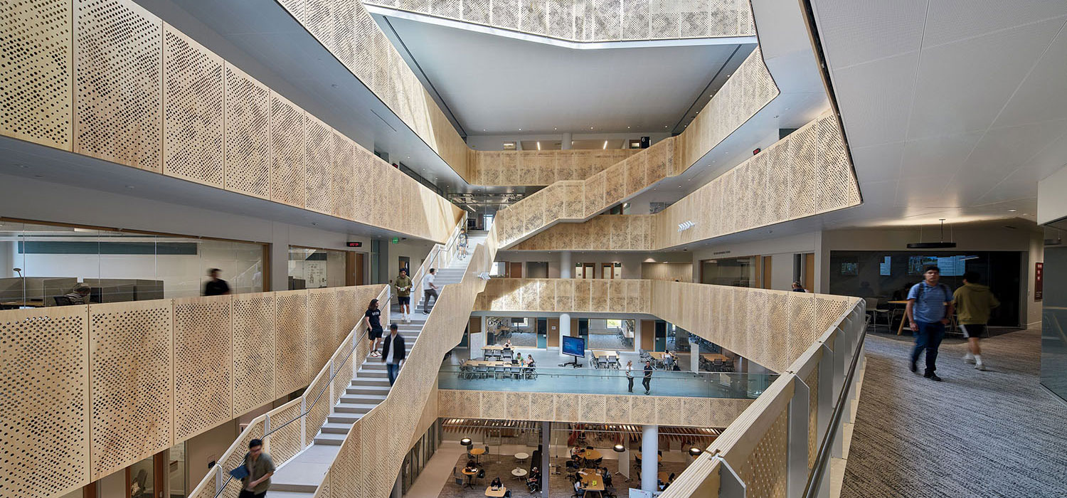 Open, modern building interior with multiple floors, angular staircases, perforated metal railings, and people walking or sitting in common areas.