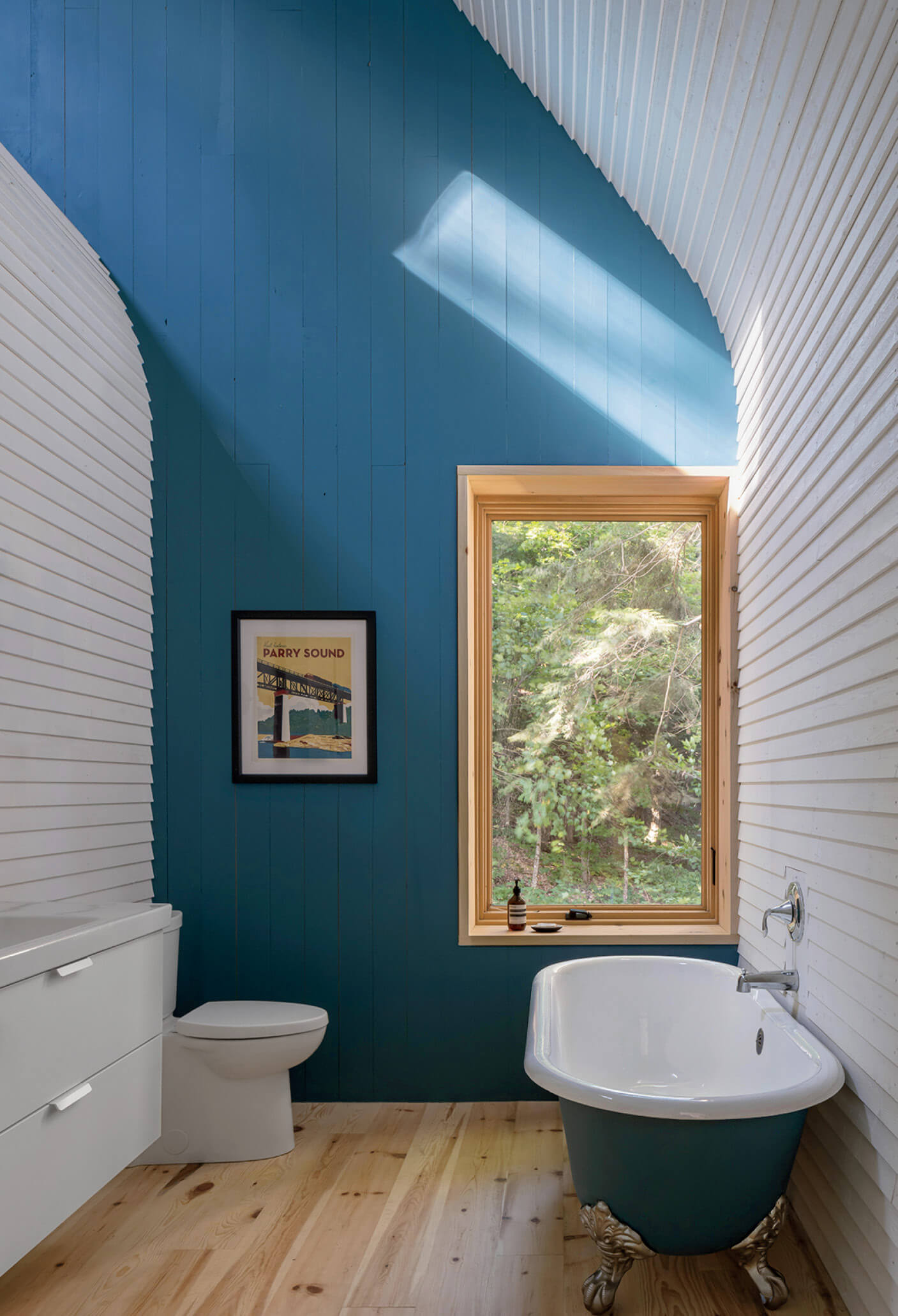 Modern bathroom with blue and white curved walls, a wooden floor, freestanding tub, toilet, framed art, and a window with greenery outside. Sunlight streams in through the window.
