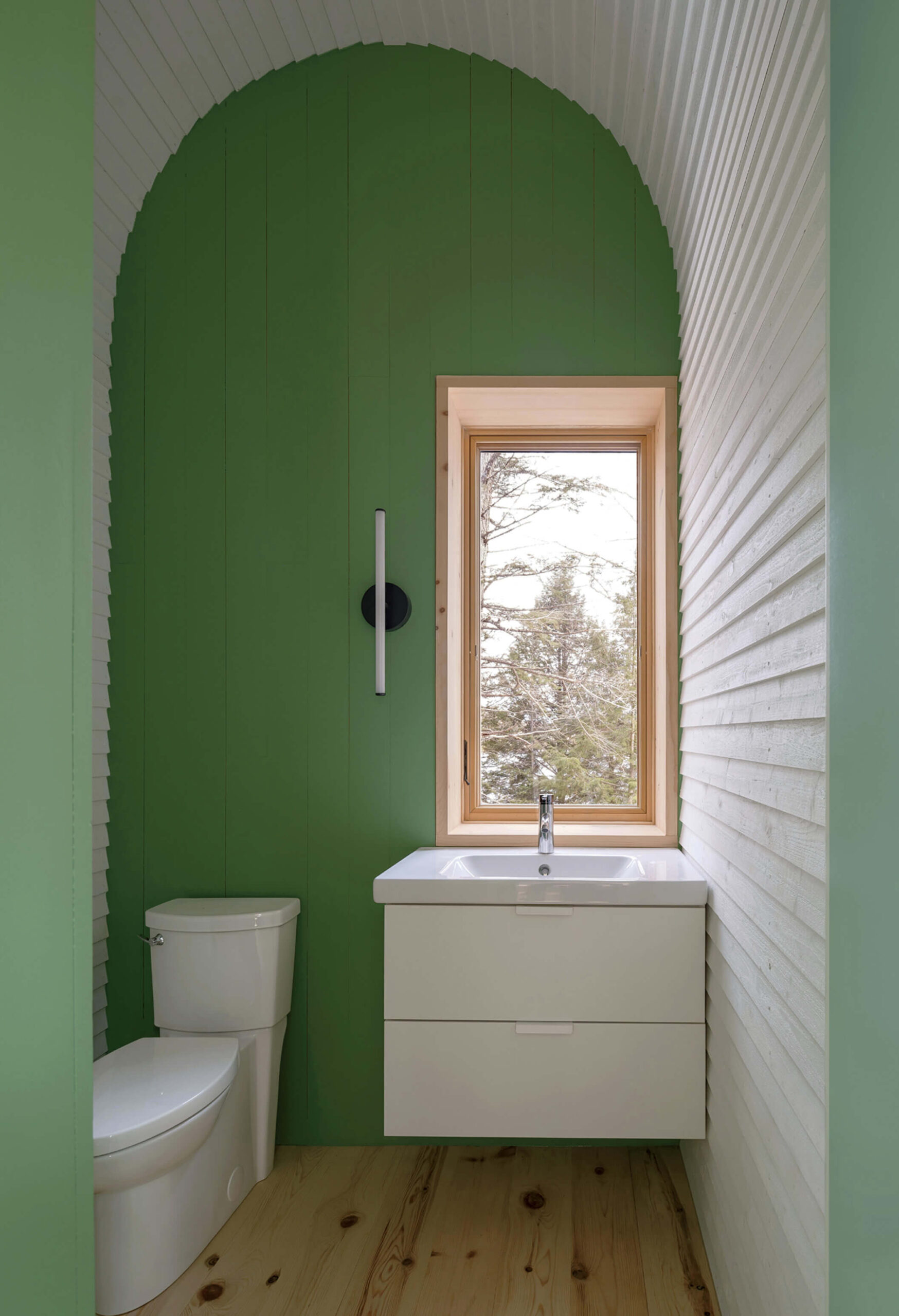 A small bathroom with green and white arched walls, a toilet, a white sink cabinet, a vertical light, and a window showing trees outside.