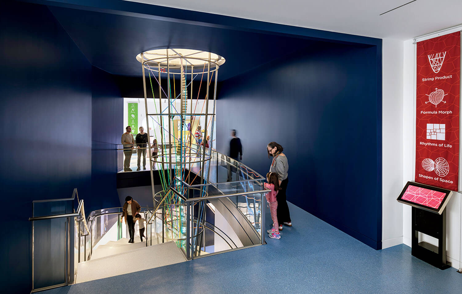 People stand and walk near a staircase with a modern railing and vertical colored rods, under a circular skylight, in a brightly lit, blue-walled interior space with educational displays.