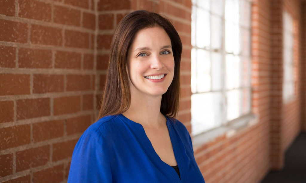 Woman with long brown hair in a blue blouse stands indoors, smiling, with a brick wall and large windows in the background.