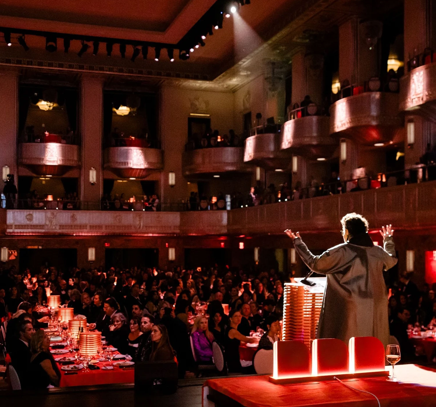 A speaker addresses a large audience seated at banquet tables in an ornate, well-lit event hall with balconies.