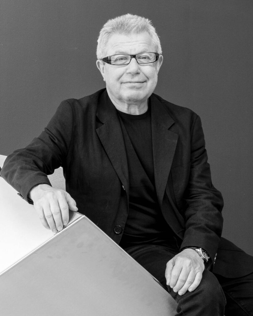 A man with short gray hair and glasses sits on a geometric metal structure, wearing a black suit and shirt, against a plain dark background.