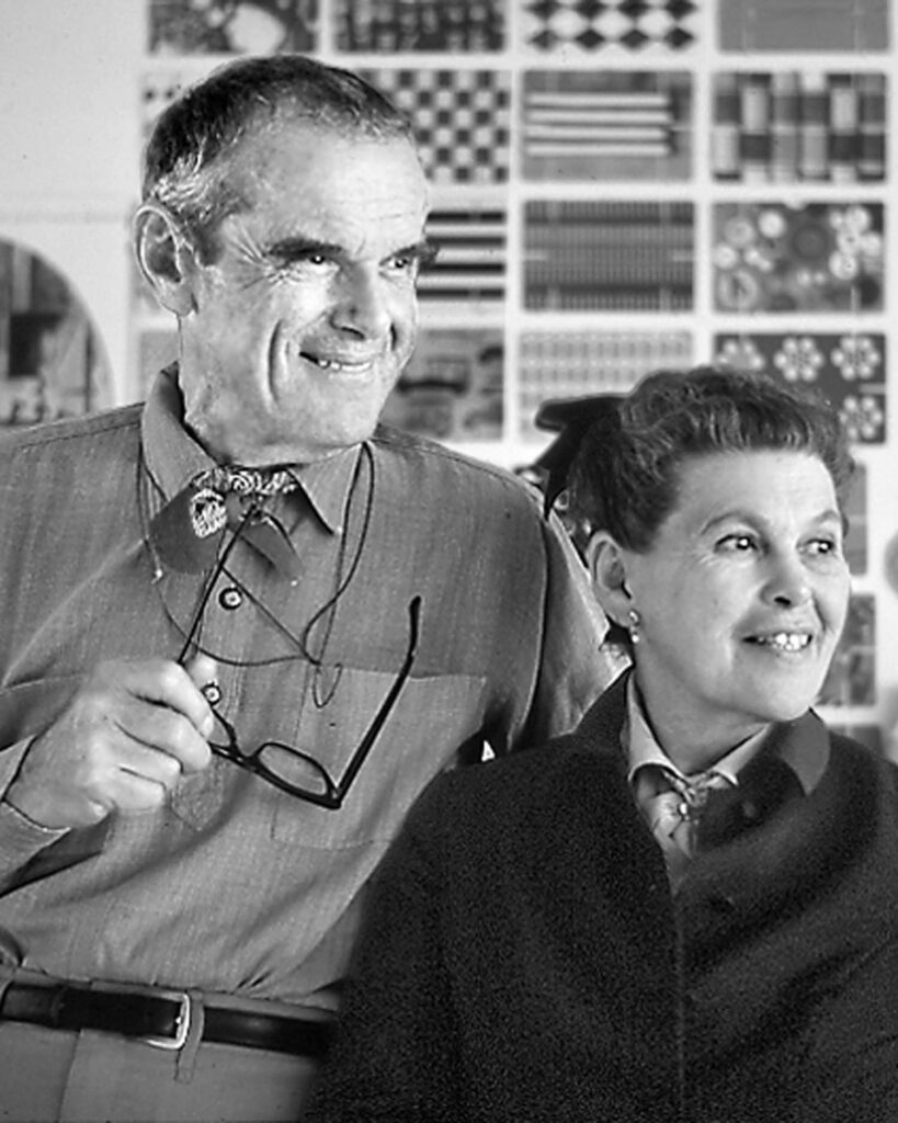 A man holding glasses and a woman stand together, smiling. The background features a wall covered with geometric art prints.