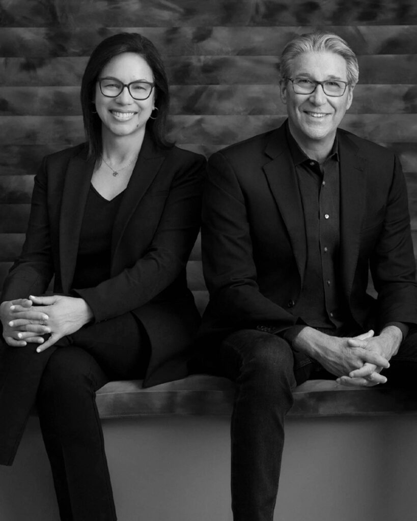 Two people wearing dark suits sit side by side on a bench, smiling at the camera. The image is in black and white with a textured background.