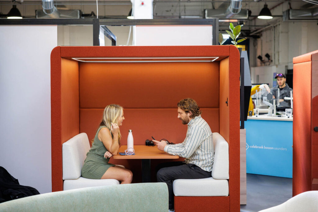 Two people sit across from each other in an orange office pod, talking. A water bottle is on the table. Office equipment and other people are visible in the background.
