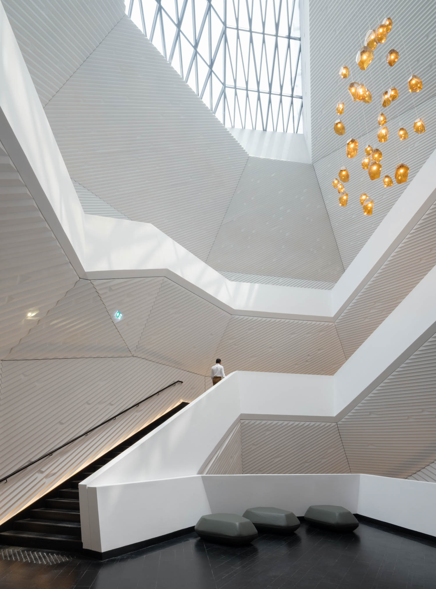A person ascends a wide staircase in a modern, angular building with geometric white walls and a glass ceiling. Clustered hanging lights are suspended from the upper right side.