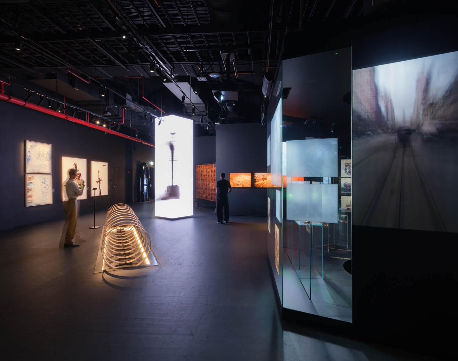 Dimly lit modern gallery with illuminated displays, abstract sculptures, and digital art projections; two people observe the exhibits.