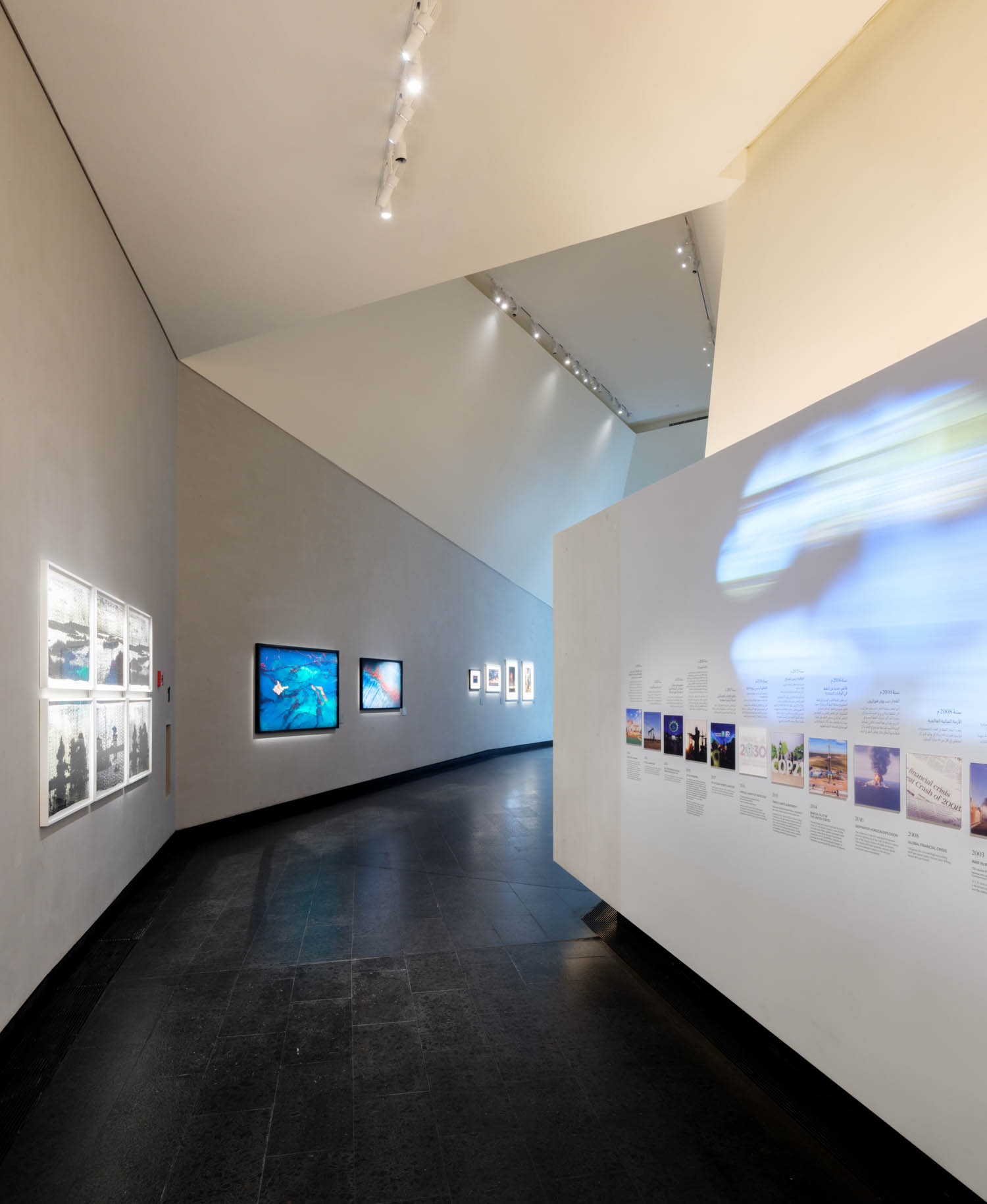 A modern museum gallery with art displays on angled white walls and illuminated photos and text panels along a dark floor.