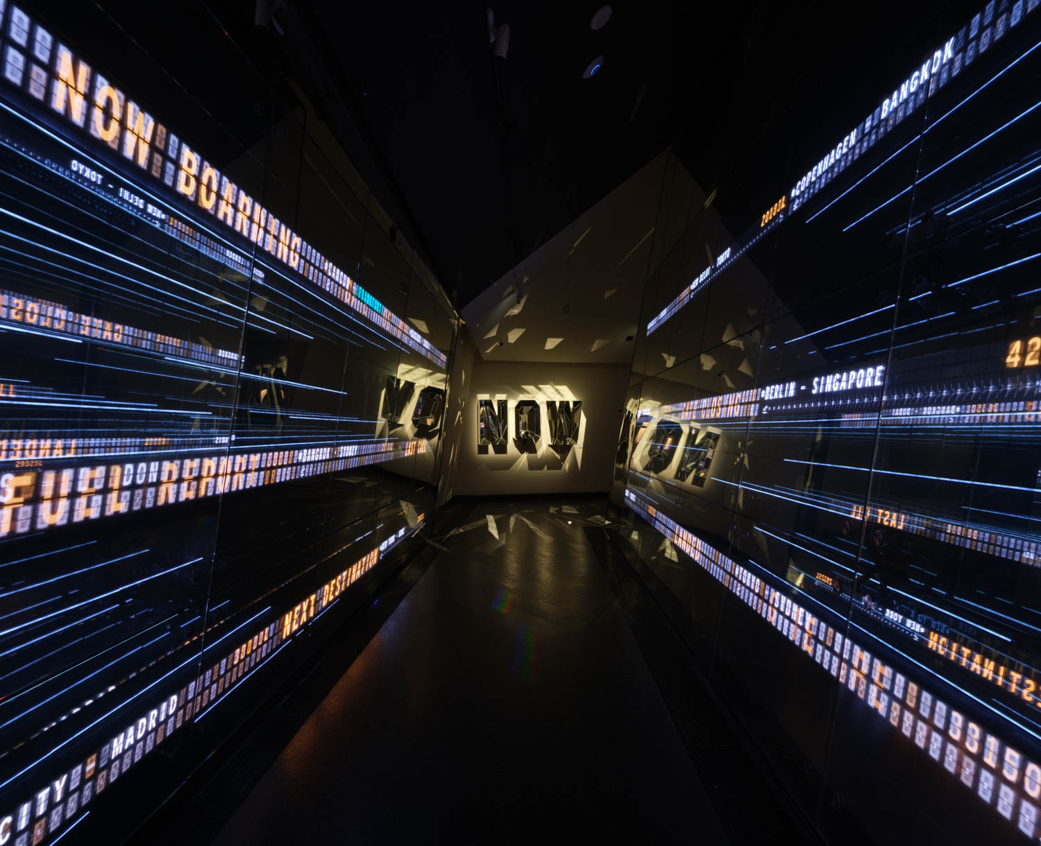 A dark hallway with mirrored digital displays showing scrolling text and bright “NOW” signage at the end.