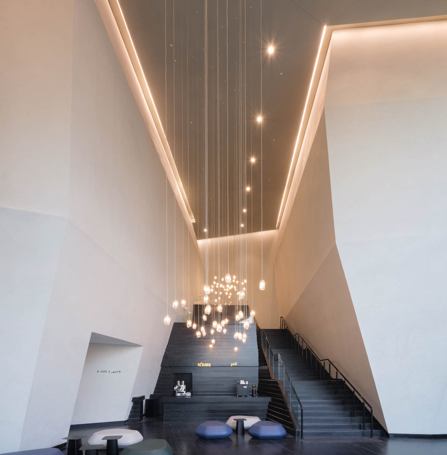 Modern lobby with high ceilings, geometric white walls, a cluster of hanging pendant lights, wide dark stairs, and minimalist seating with round ottomans.