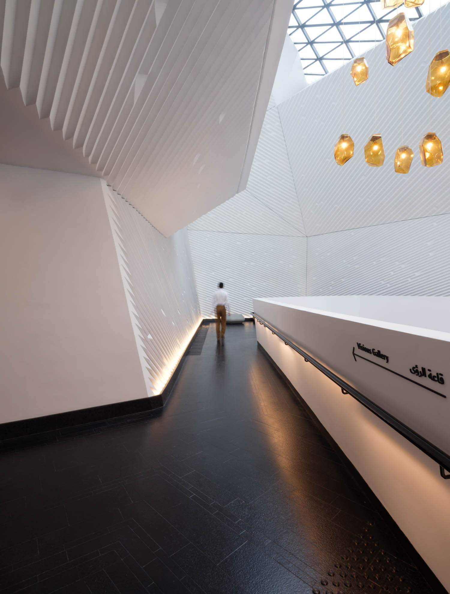 A person walks along a modern, white, angular corridor with dark flooring, geometric walls, and hanging amber lights.