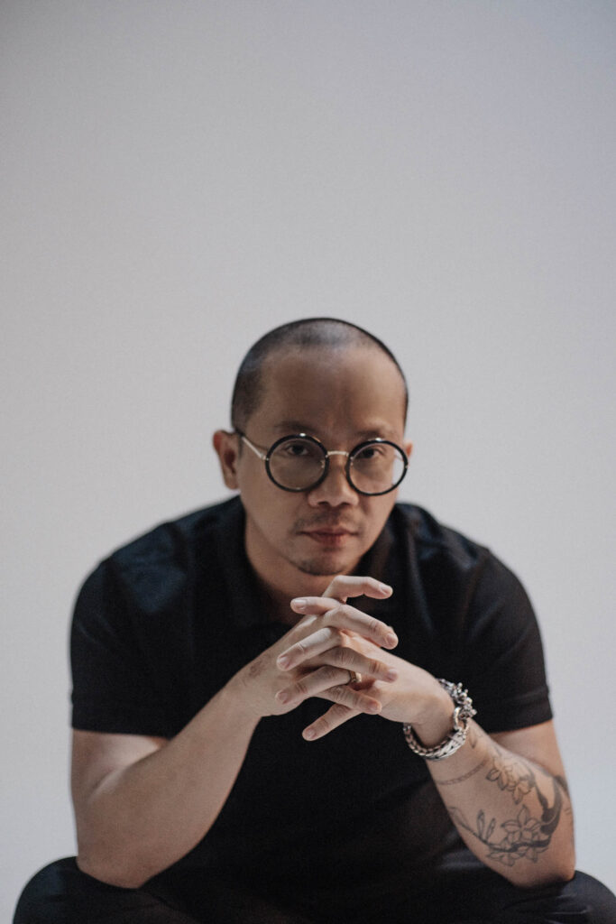 A person with glasses and a tattoo on their left forearm sits with hands clasped, wearing a black shirt, against a plain light background.