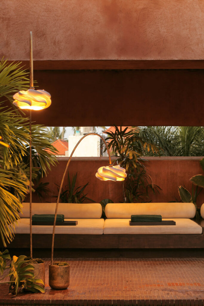 Outdoor lounge area with modern lighting, cushioned seating, potted plants, tile flooring, and a terracotta wall.