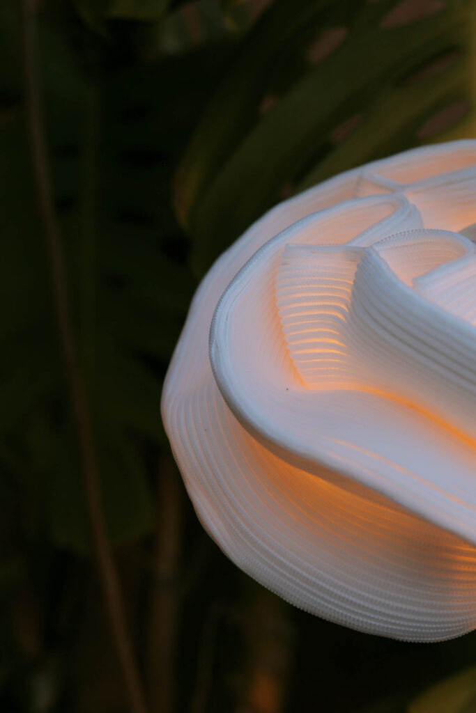 Close-up of a white, sculptural lamp with a soft orange glow, set against a dark background with green leaves partially visible.