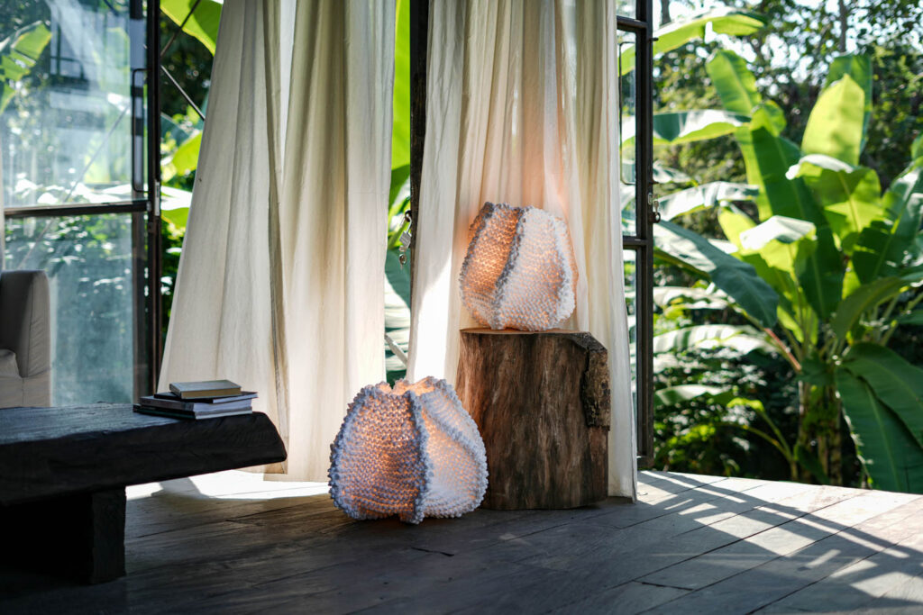 A sunlit room with sheer curtains, two textured lamps on a tree stump, books on a dark table, and lush green plants outside.