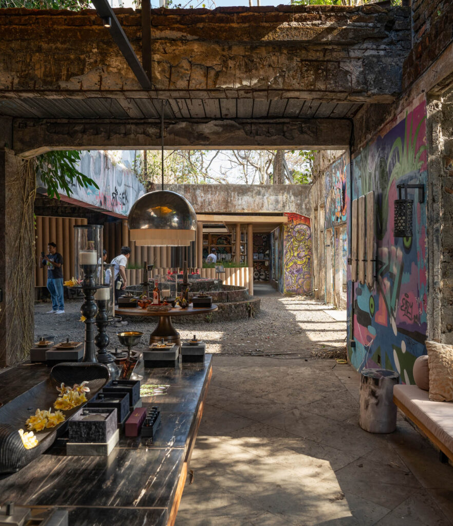 Abandoned building repurposed as a café with graffiti-covered walls, exposed brick, and people in the background. Tables display food and decorations. Sunlight streams through openings.