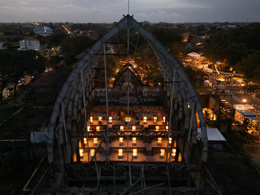 Aerial view of a partially roofless, abandoned building repurposed with lit tables inside for an evening event, surrounded by trees and city lights at dusk.