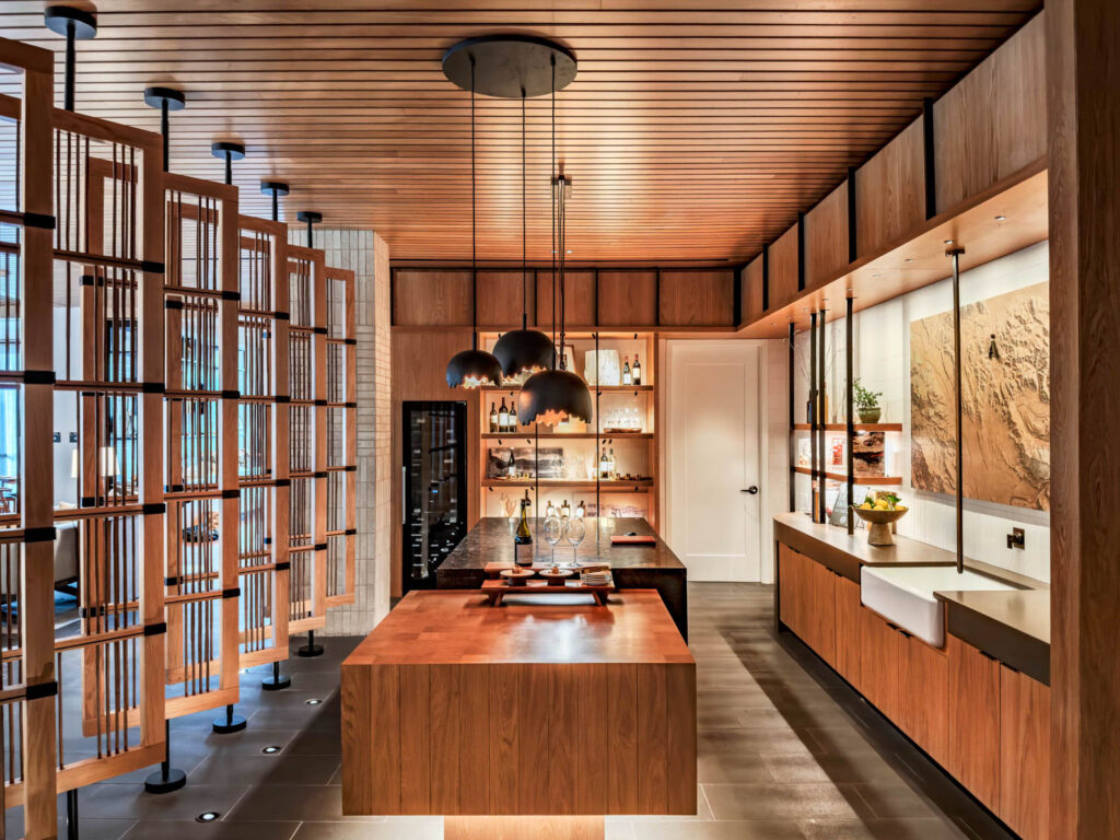 Modern kitchen with wood cabinetry, a large island, pendant lighting, open shelving, and a decorative wood slat partition.