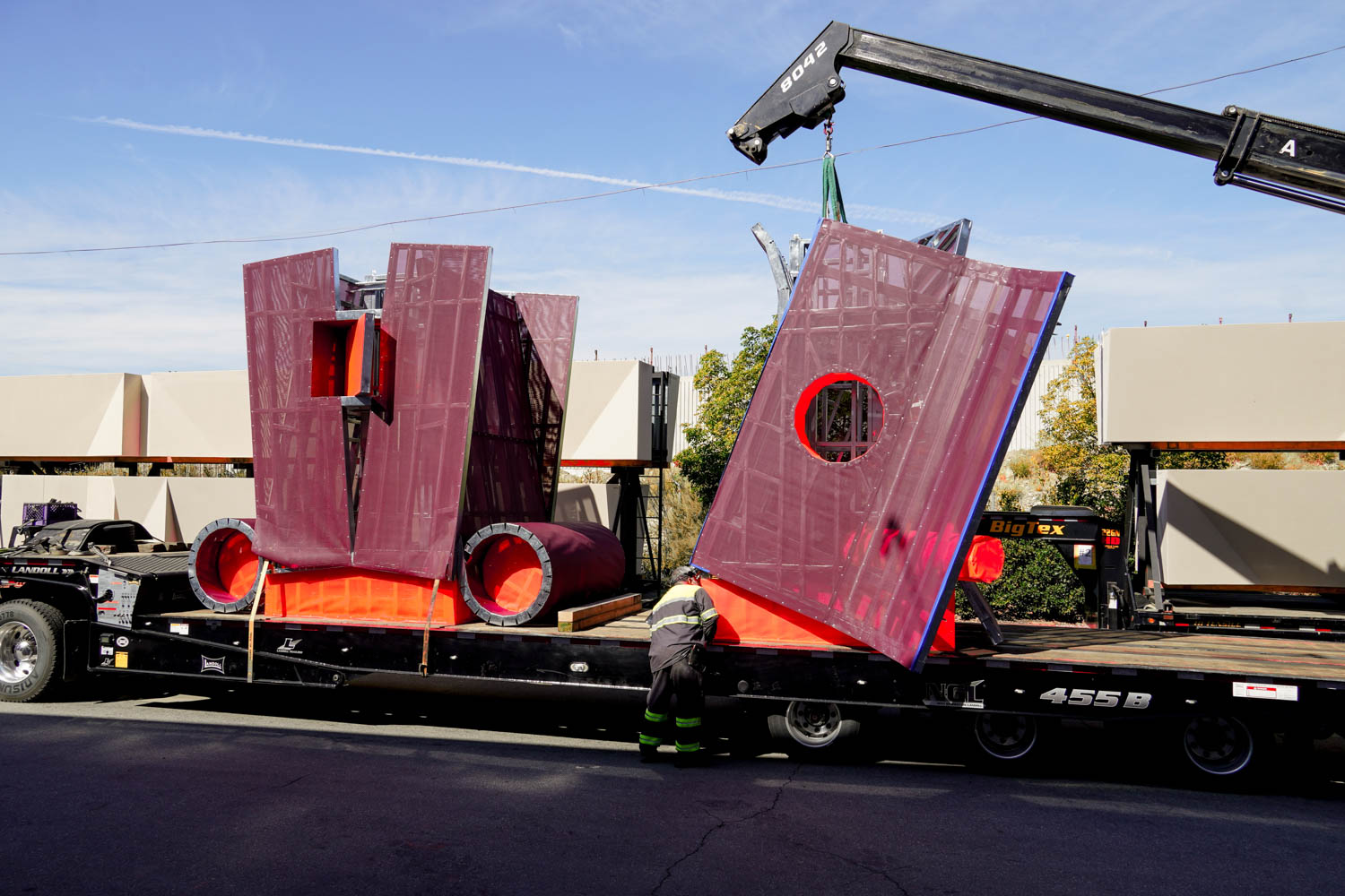 A worker stands beside a flatbed truck carrying large, red and purple geometric structures, while a crane lifts one section.