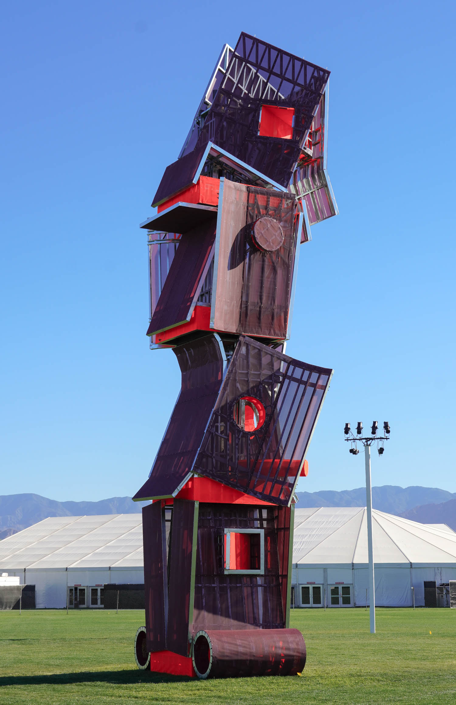 A tall outdoor sculpture made of stacked, angular, reddish metal and glass structures stands on grass, with a tent, mountains, and blue sky in the background.