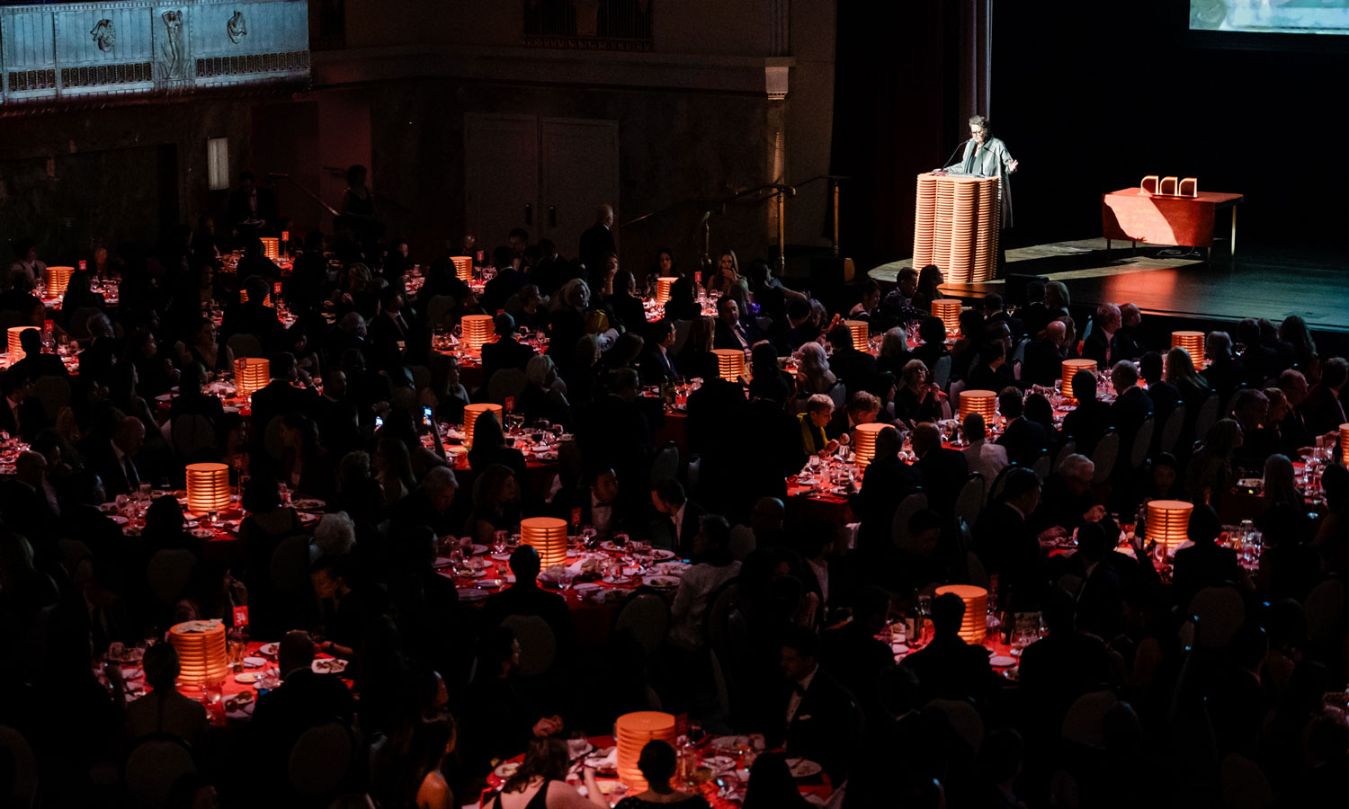 A speaker stands at a podium on stage addressing a large audience seated at round tables with orange lamps during a formal event in a dimly lit hall.
