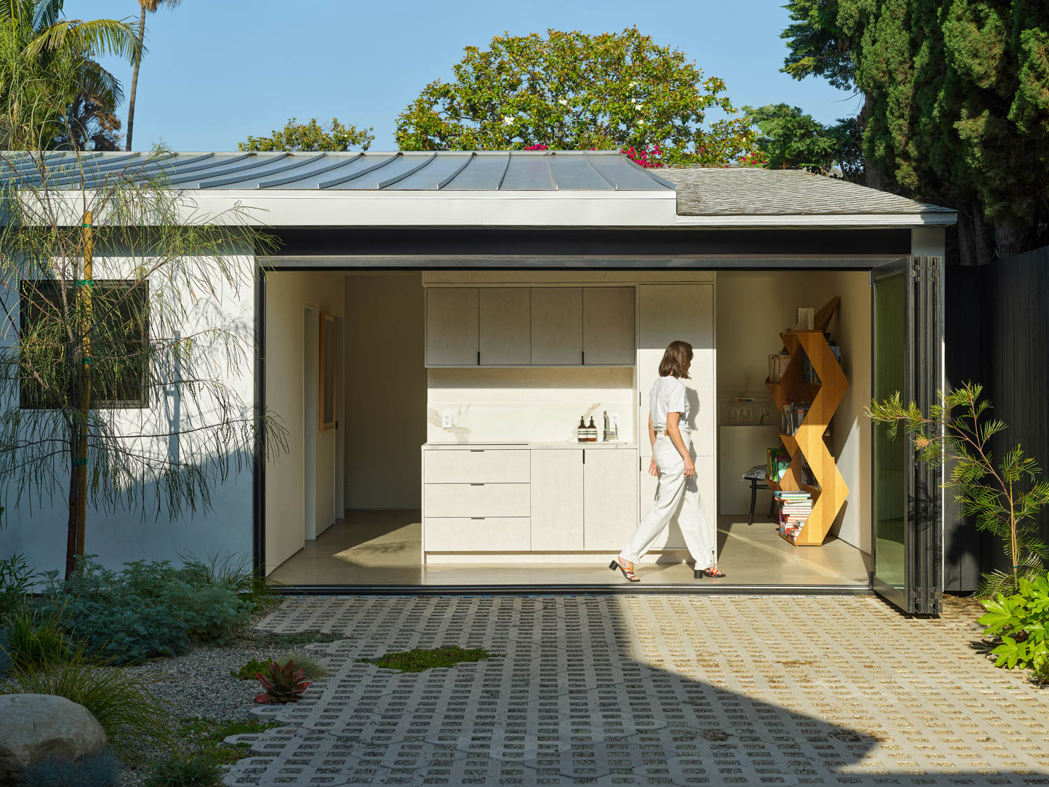 A person in white clothing walks inside a modern garage converted into a kitchen; plants and trees surround the exterior under clear daylight.