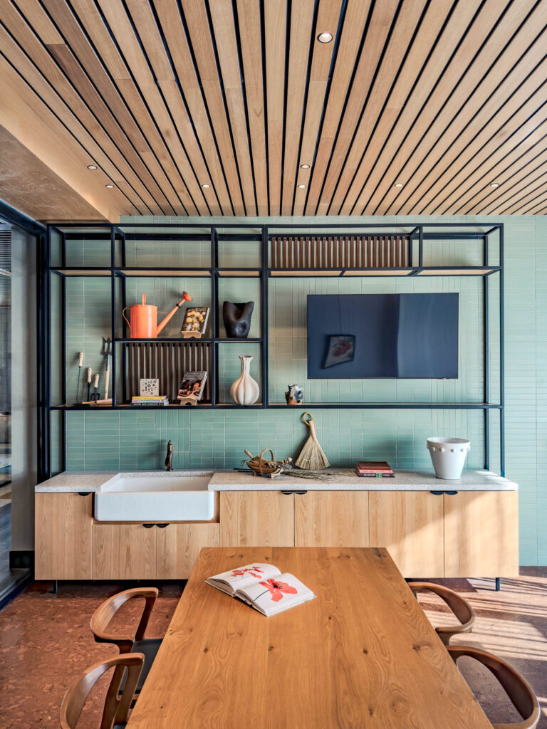 Modern kitchen with open shelving, sink, decorative items, flat-screen TV, wooden table, and chairs under a slatted wood ceiling; a book is open on the table.