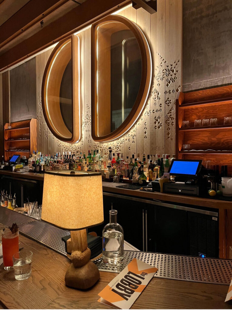 A dimly lit bar with a large round mirror, shelves of liquor bottles, a menu labeled “Lobby,” a drink, a lamp, and a bottle of water on the counter.