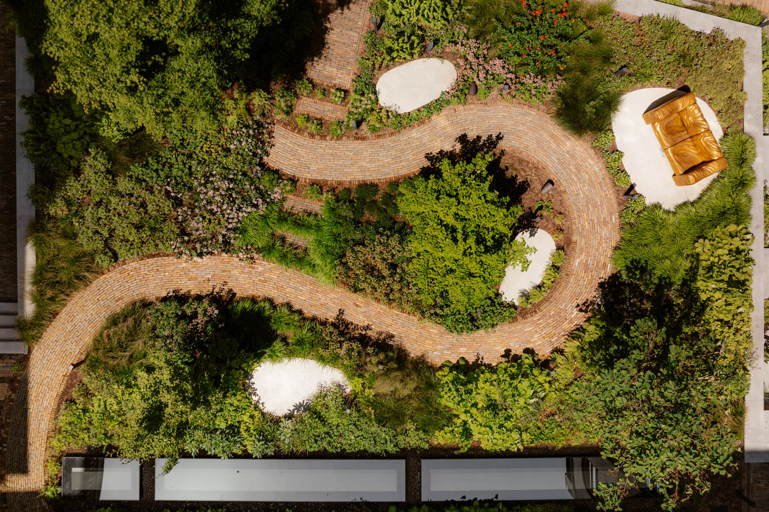 A winding brick path curves through a green garden with trees, plants, white gravel patches, and a yellow bench.