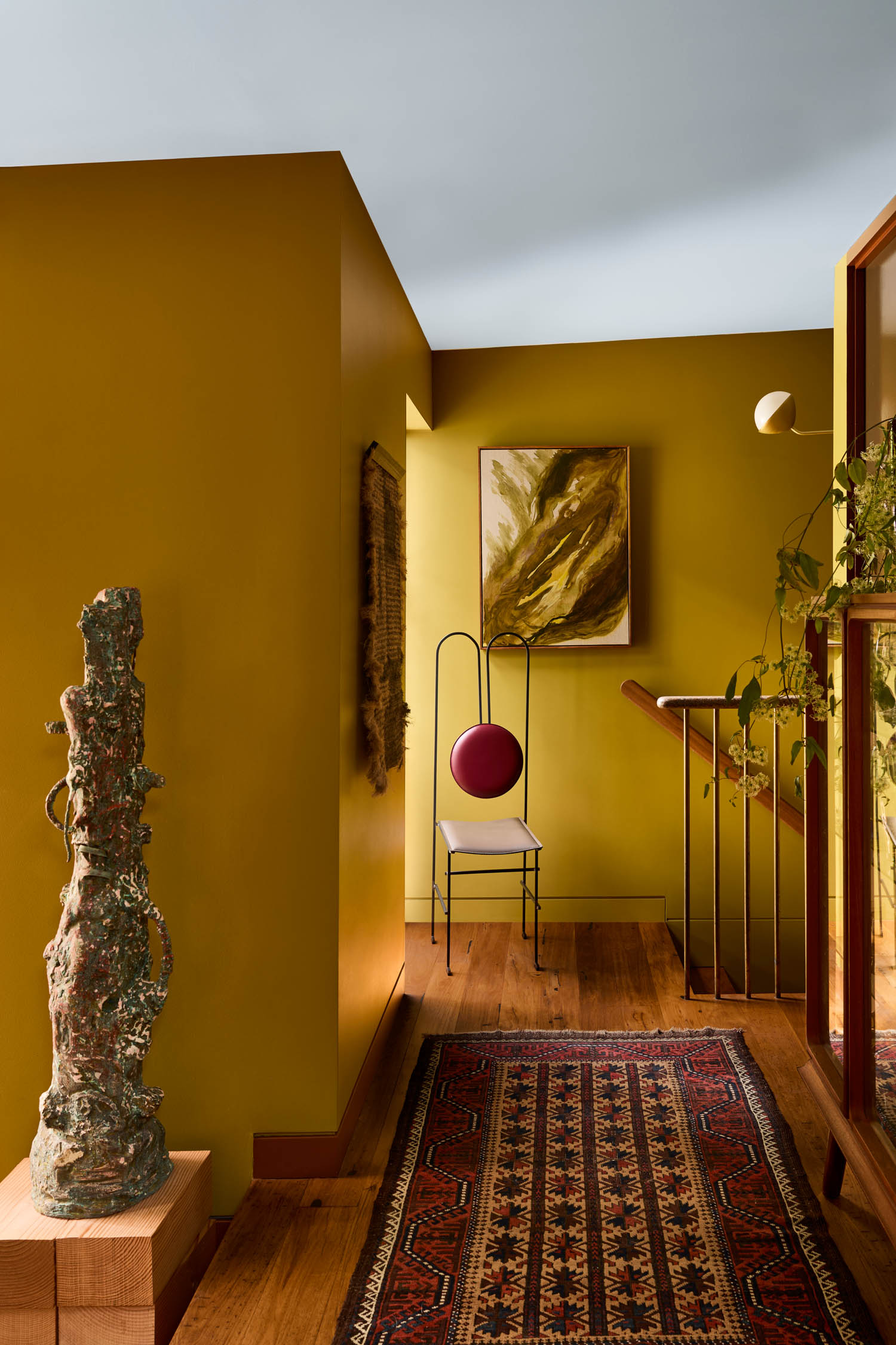 A hallway with mustard-yellow walls, abstract art, a tall sculpture, a black metal chair with a red cushion, a patterned rug, and wooden floors.