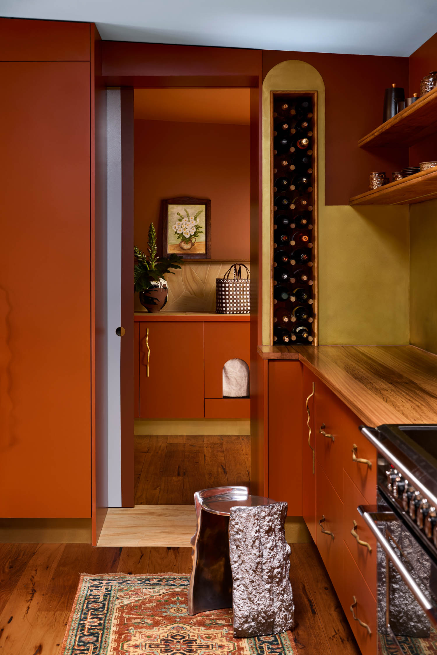Modern kitchen with burnt orange cabinets, wooden countertop, built-in wine rack, open door to a laundry area, and a metallic stool on a patterned rug.