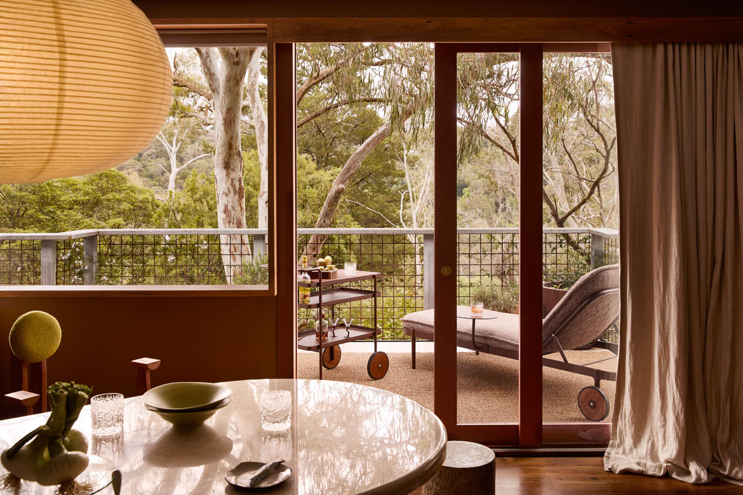 A dining area with a round table overlooks a balcony with a lounge chair and a drink cart, framed by glass doors and surrounded by greenery outside.