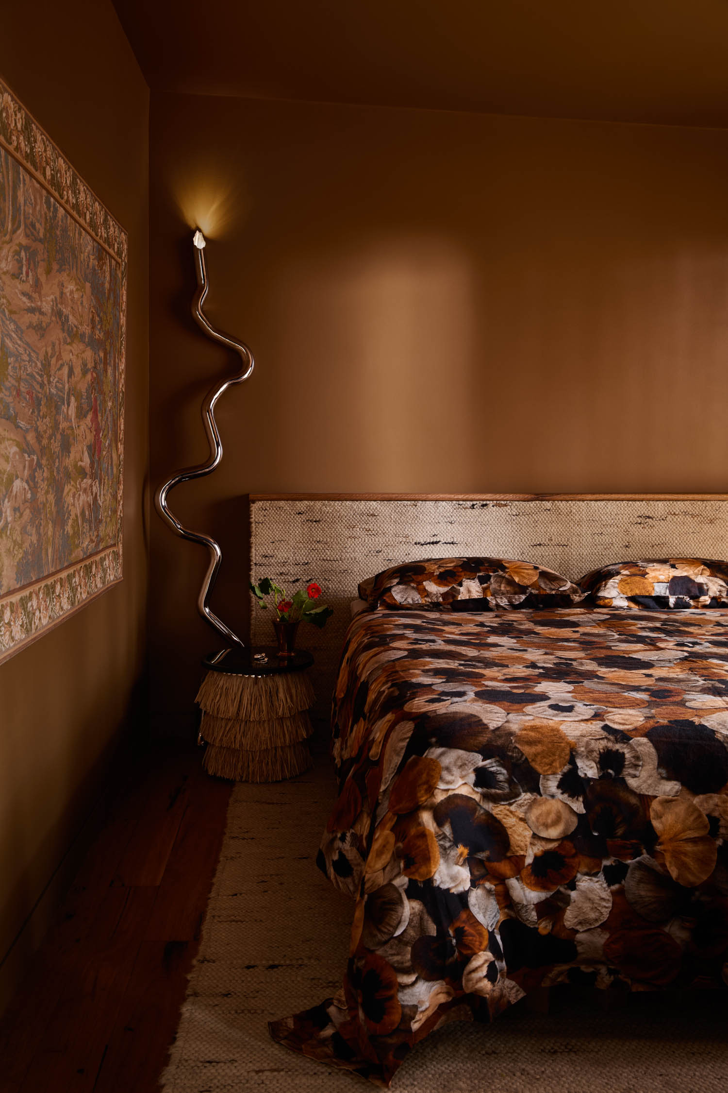 A bedroom with brown walls, a textured headboard, a floral-patterned bedspread, a wavy wall lamp, and a small round bedside table with a vase of flowers.