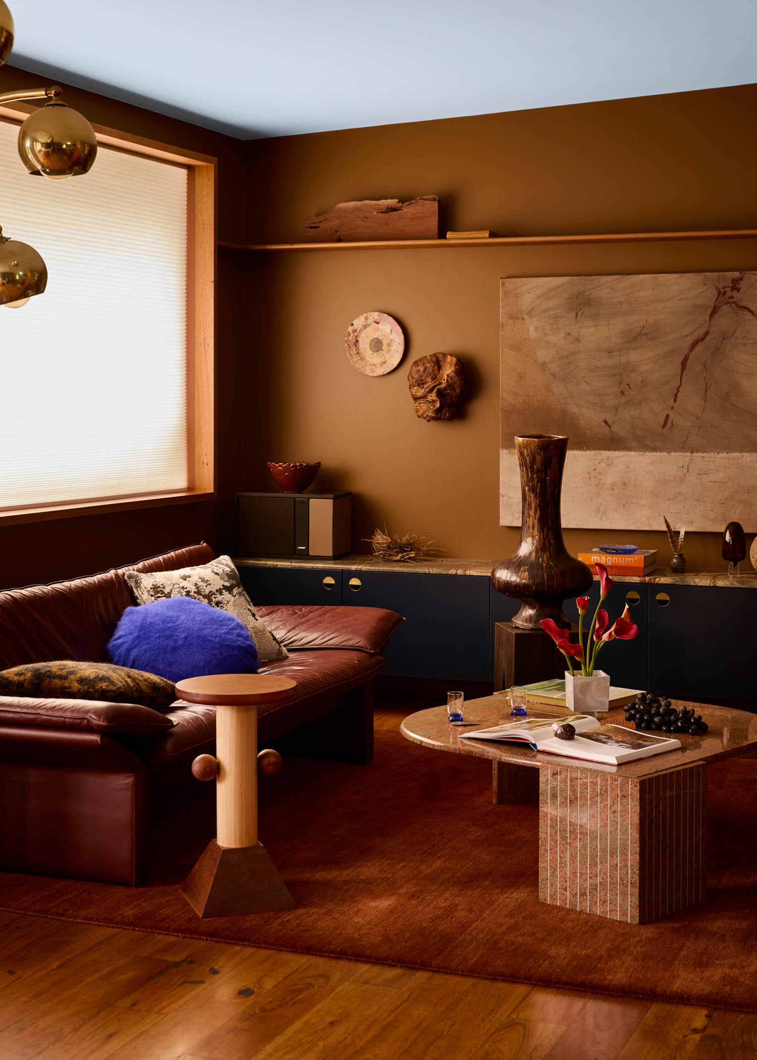 A cozy living room with brown walls, a leather sofa, modern coffee table, eclectic decor, a vase with flowers, and abstract art on display.