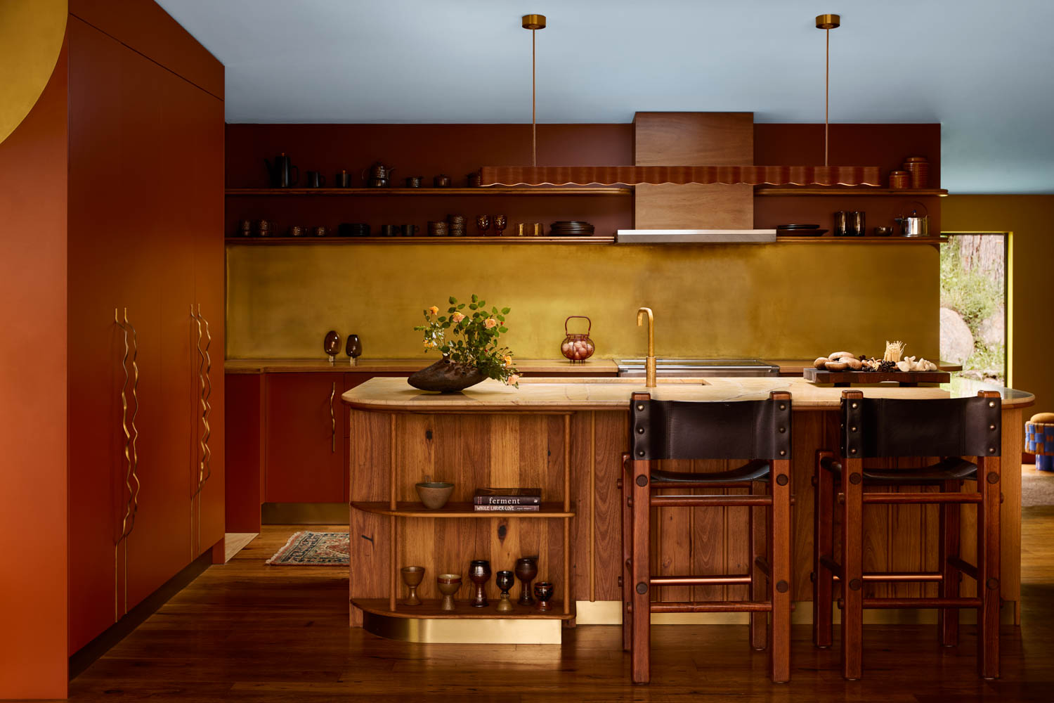 Kitchen with warm brown and gold tones, wood island with shelves, two high chairs, open shelving, and various kitchenware arranged neatly.