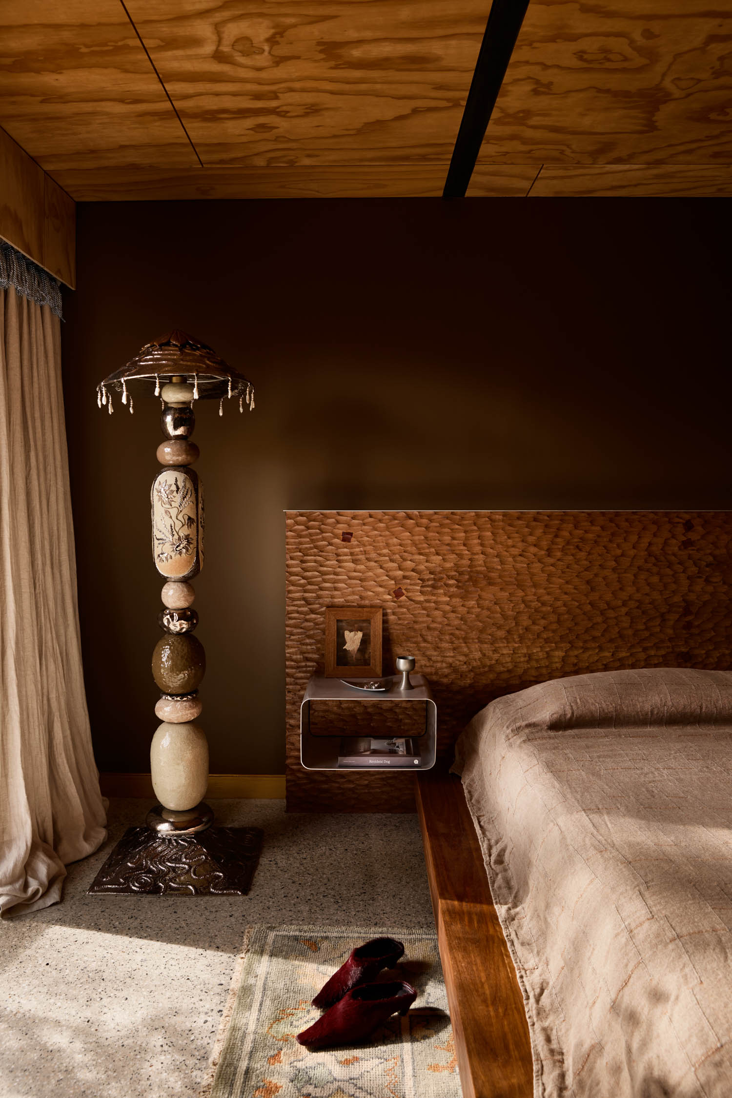 A bedroom with a wooden bed, brown bedding, a textured headboard, an ornate floor lamp, a small bedside table with a framed photo, and slippers on a rug.