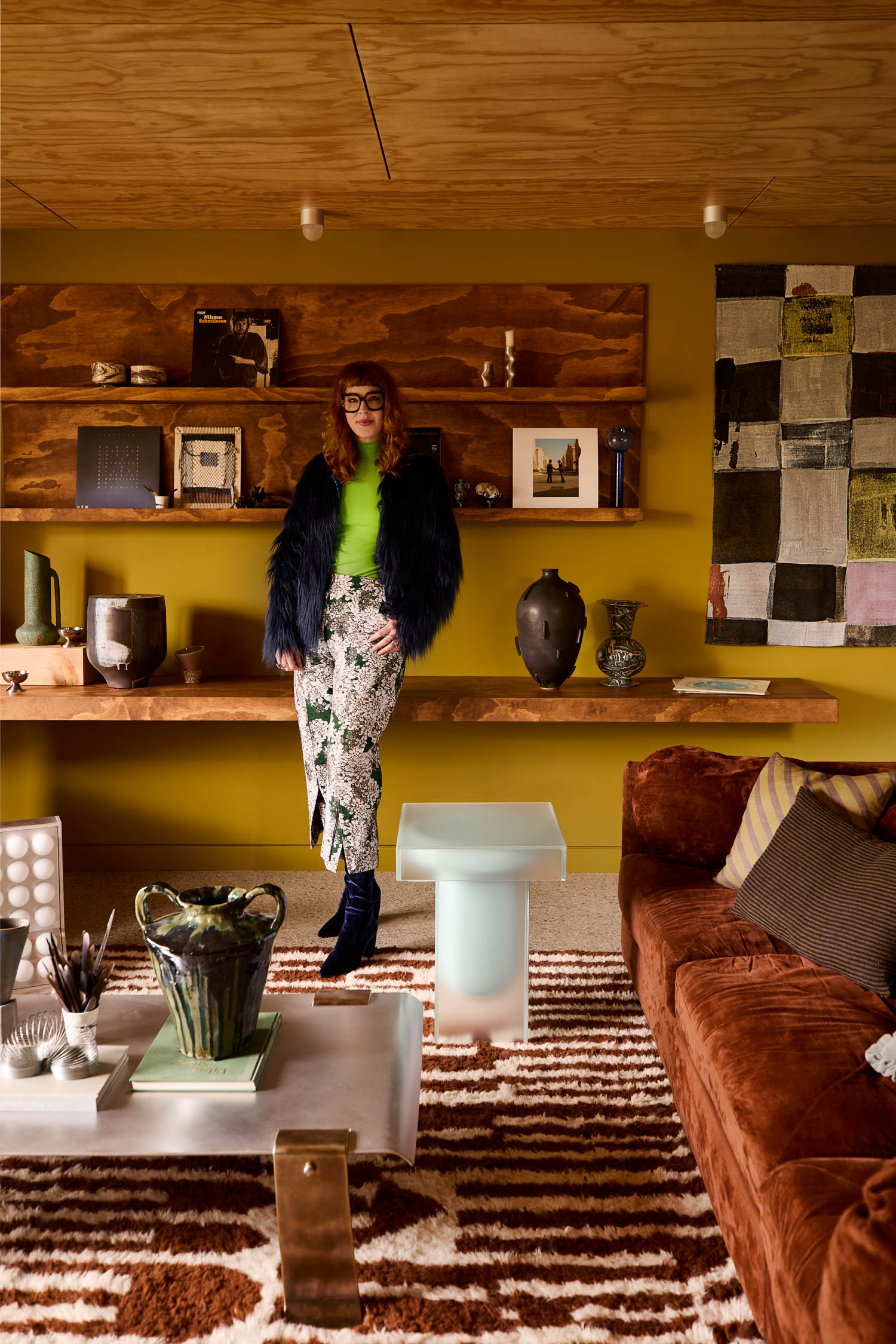 A woman in a neon green top and patterned skirt stands in a stylish living room with earth-toned walls, floating wooden shelves, modern decor, and a rust-colored sofa.
