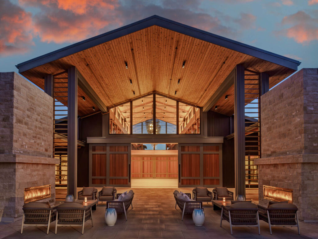 Outdoor seating area with modern chairs and tables in front of a large building with wood and stone accents, warm lighting, and a pitched roof at sunset.