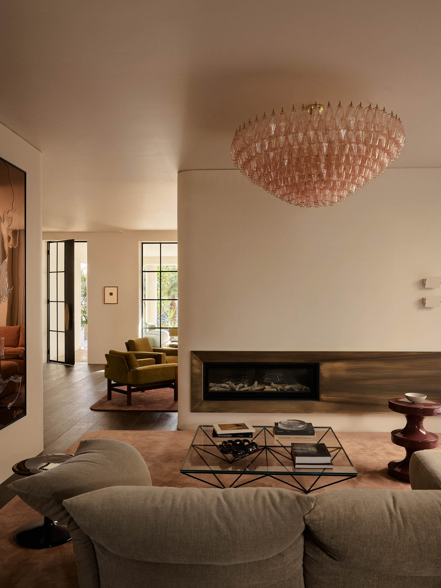 A modern living room with a beige sofa, geometric glass coffee table, large pink chandelier, built-in fireplace, and green armchair near a windowed doorway.