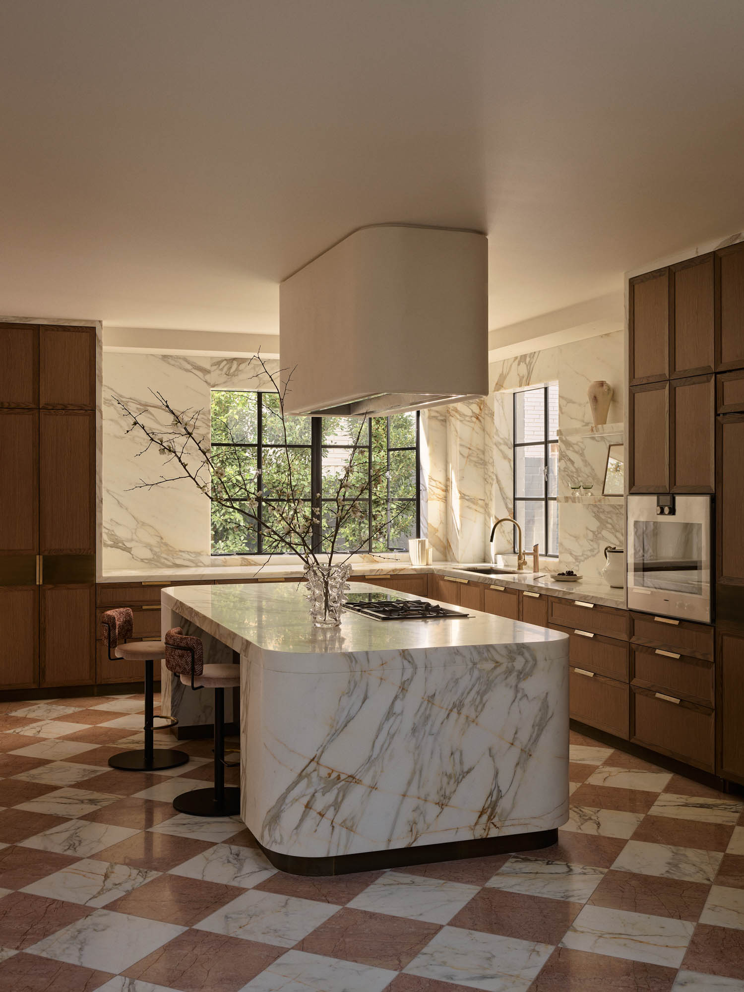 Modern kitchen with marble island, brown cabinets, built-in appliances, checkerboard tile floor, large windows, and a glass vase with branches on the counter.