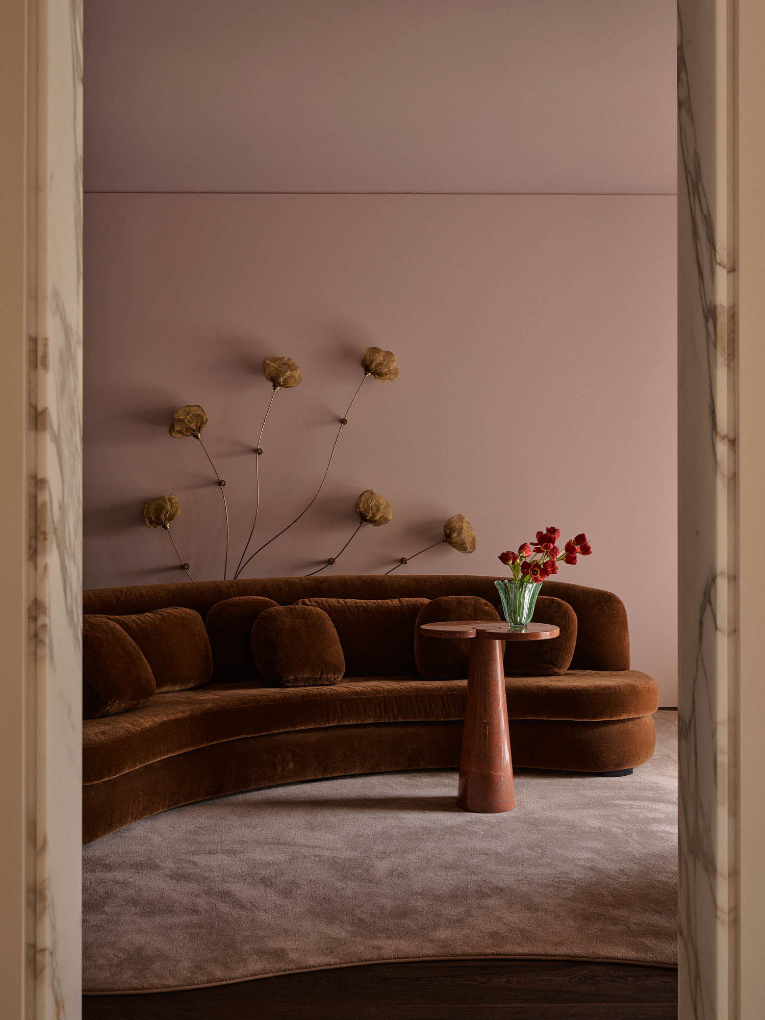 A curved brown velvet sofa, round side table with red flowers, and wall art of golden flowers in a minimalist room with pink walls and a light rug.